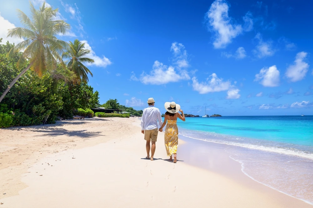 A couple walking along the white sand beach of Antigua between the turquoise ocean and green trees.