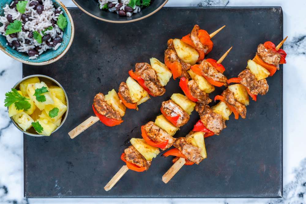 Three jerk chicken skewers with pineapple and pepper on a black slate.