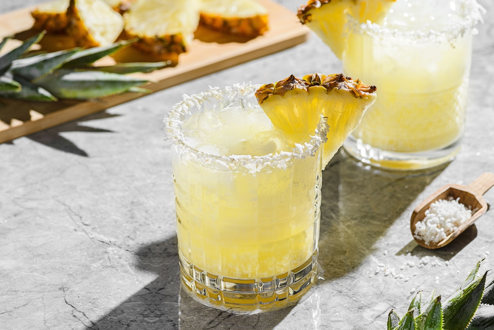 Two glasses of pina colada with pineapple as a garnish and a chopping board with pineapple in the distance.