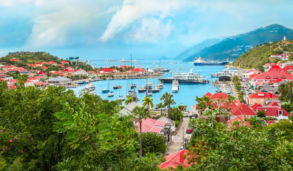 Air view of St Barts' harbour with a superyacht on the bright blue water and red roofed buildings.