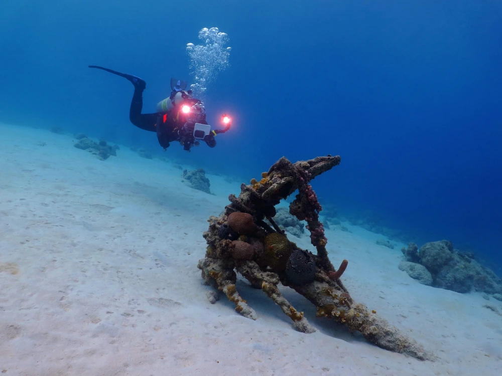 A man diving underwater approaches a shipwrecked anchor.