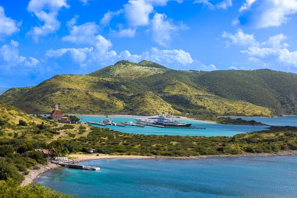 A yacht in the harbour of St Kitts and Nevis on bright blue water beside tall green covered hills.