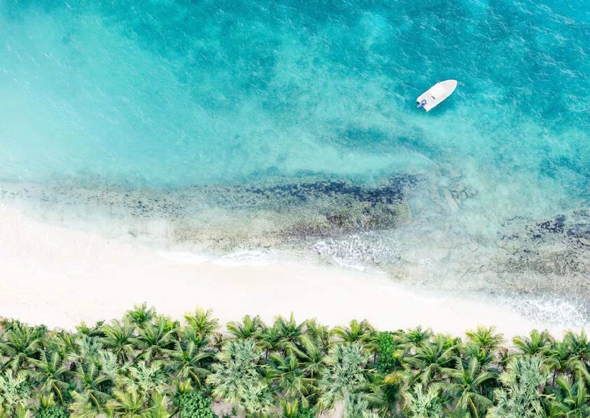 Birdseye view of a yacht off the coast of St Martin in bright blue waters besides white beaches and a line of tropical trees.