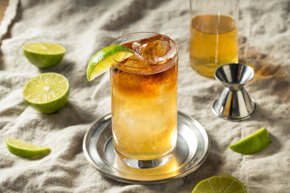 Spiced rum cocktail in a glass with ice and a lime wedge on a small silver dish beside cut-up lime, a jigger measurer and a glass of mixed.