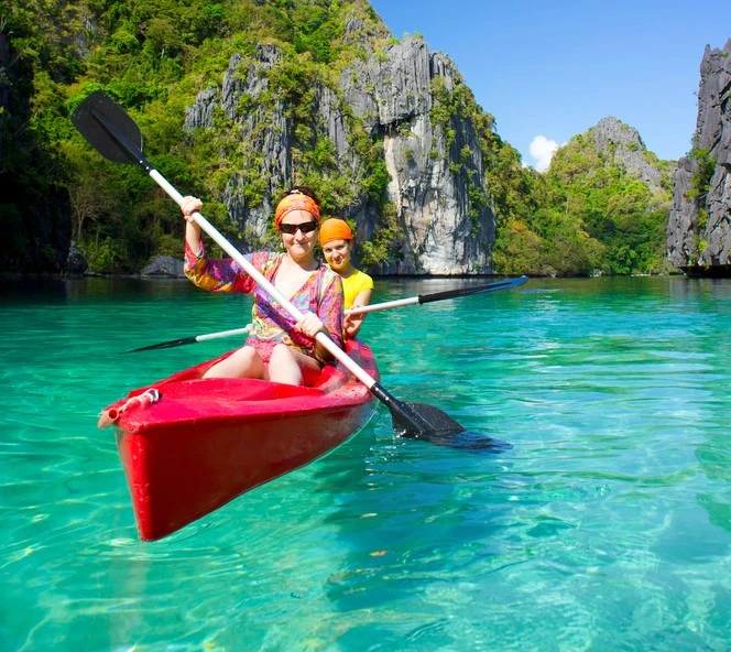 Two women in a red kayak, kayaking on bright blue water with a rockface covered in green.