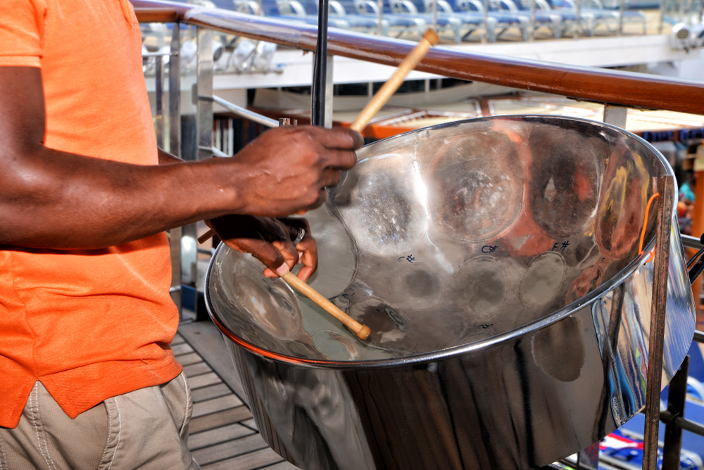 The hands of a black man in an orange shirt playing a steel drum with wooden drunsticks.