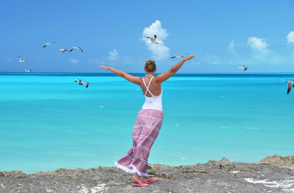 A woman with arms outstretched, staring at birds flying over turquoise water.