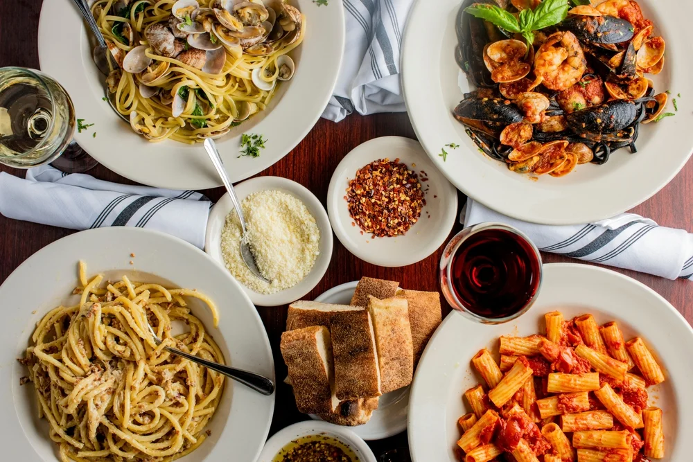 A spread of Italian food and drink with multiple pasta dishes and a bowl of bread, parmesan, and chilli flakes in the middle of the table.