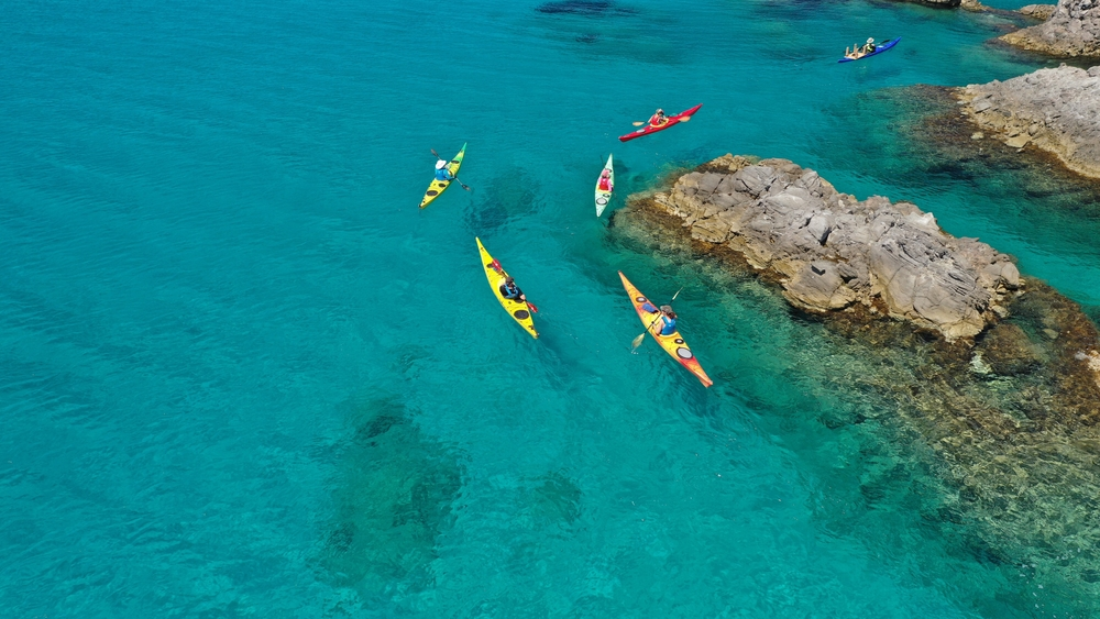 Five kayaks moving through turquoise waters around Greek rocks.