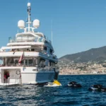 The Desamis B superyacht off the coast of land with watertoys behind it on a clear day.